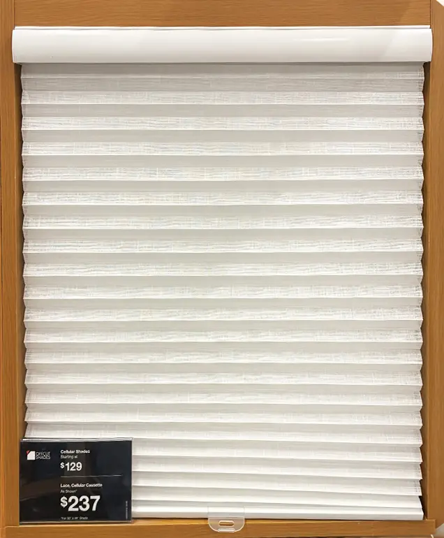 Alternative Text: White cellular honeycomb shade displayed in a wood-framed showroom cabinet with a pricing tag reading $129 and $237