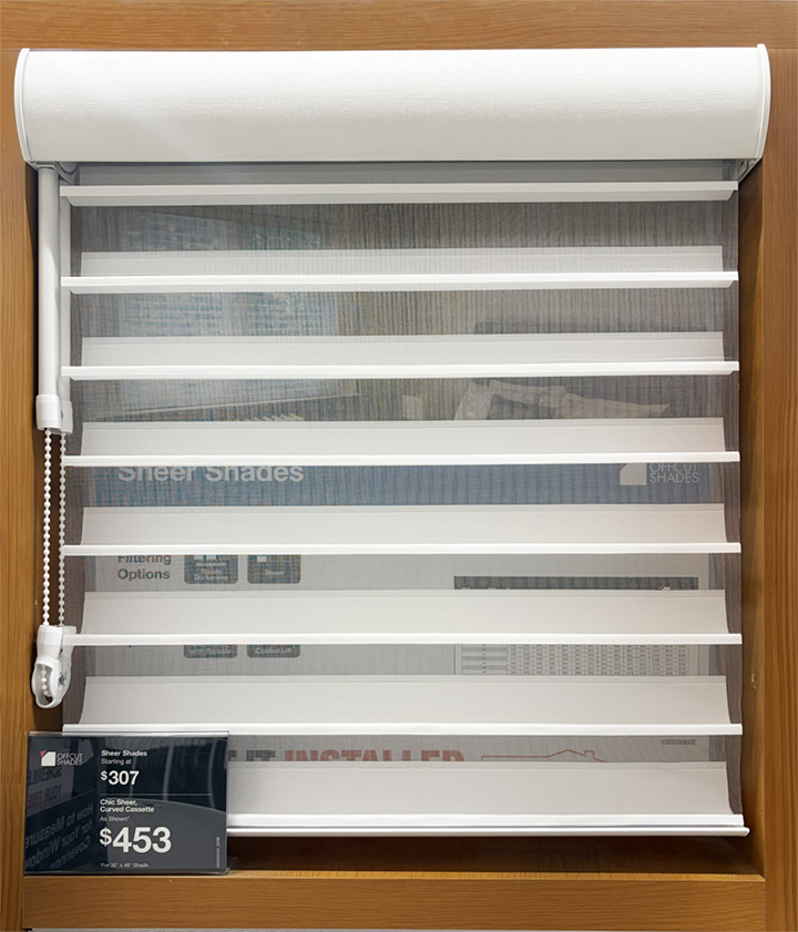 White sheer shade with horizontal fabric vanes and chain control displayed in a wood-framed showroom cabinet with a pricing tag reading $307 and $453
